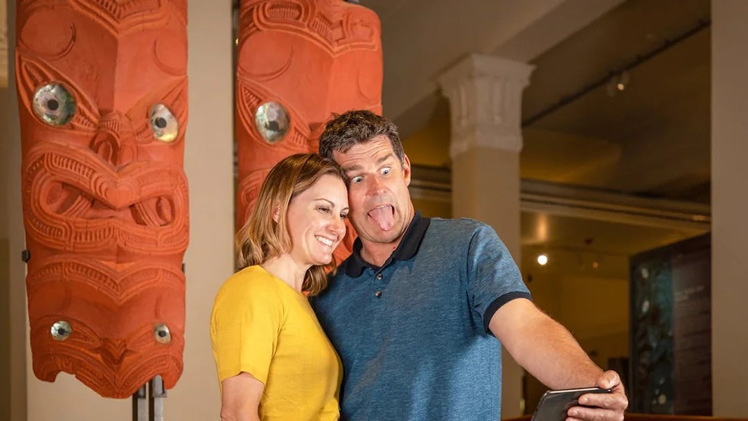 Couple taking selfie with Māori carvings at Auckland museum on hop-on-hop-off tour.