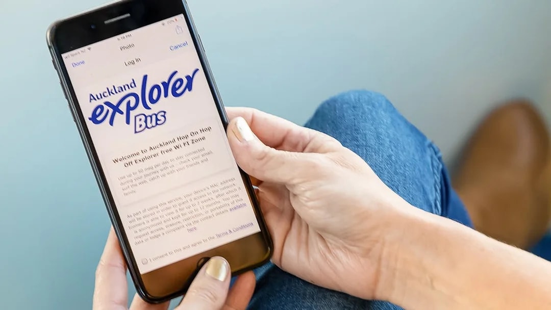 Person holding a phone displaying Auckland Explorer Bus hop-on-hop-off tour information.
