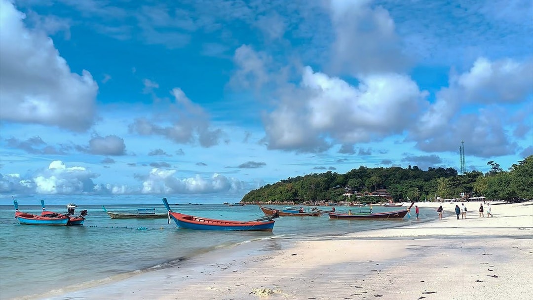 Boats anchored near the shore of Koh Lipe with tourists walking on the beach.