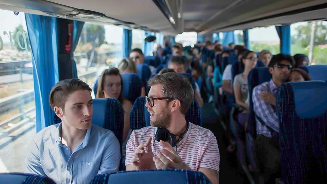 Passengers conversing on a tour bus with blue seats and curtains.
