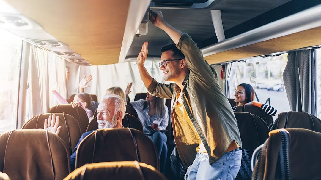 Passengers enjoying a bus ride, some standing and waving, with sunlight streaming through windows.