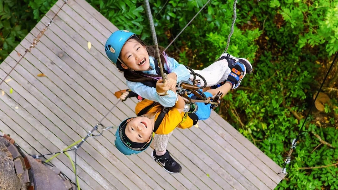 Children ziplining at Flying Hanuman, Phuket, on a wooden platform amidst lush greenery.