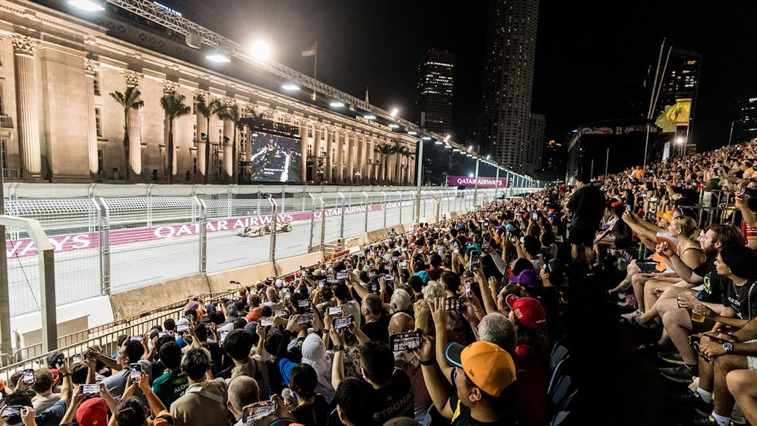 Crowd watching F1 race at night in Singapore with city skyline in background.