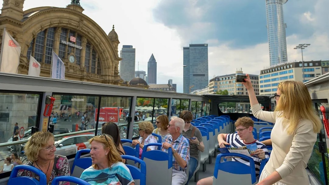 Tourists on a hop-on hop-off bus near Frankfurt Hauptbahnhof with city skyline in view.
