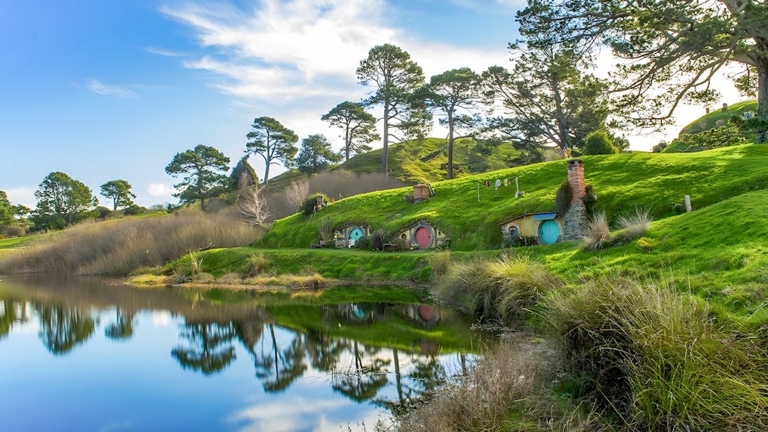 Hobbiton Movie Set with hobbit holes and lush green hills reflected in a pond, near Auckland.