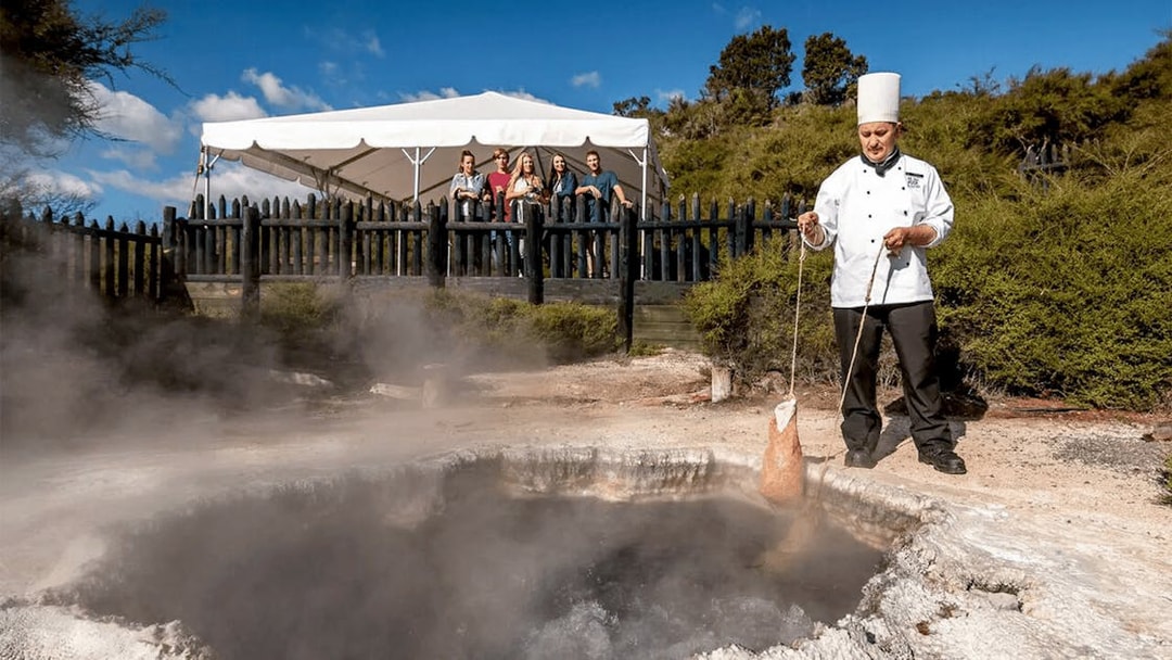 Chef demonstrating geothermal cooking at Te Puia, Rotorua, with tourists observing.