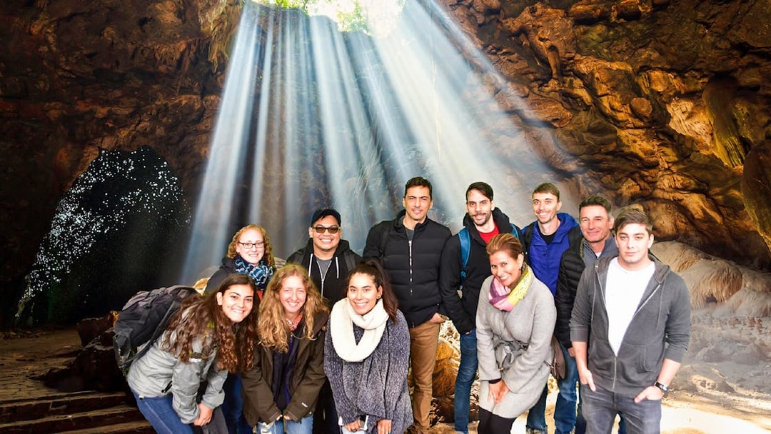 Group exploring Waitomo Glowworm Caves with light streaming through cave opening, Auckland tour.
