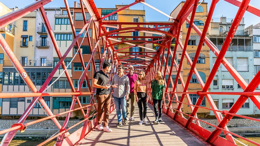 Tourists with guide on Eiffel Bridge in Girona, colorful buildings in background.