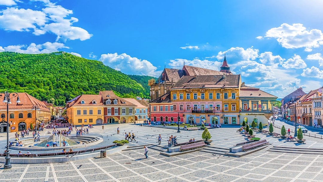 Brasov old town square with colorful buildings and people, surrounded by green hills.