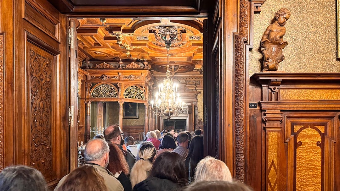 Guests touring ornate interior of Peles Castle with carved wood and chandelier.