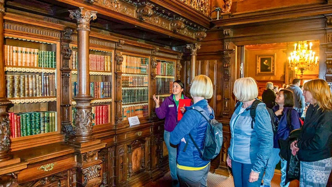 Guided tour group in Peles Castle library, Romania, viewing ornate wooden bookshelves.