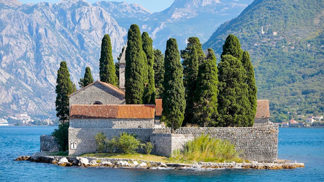 St. George Island in Perast with stone church and cypress trees against mountain backdrop.