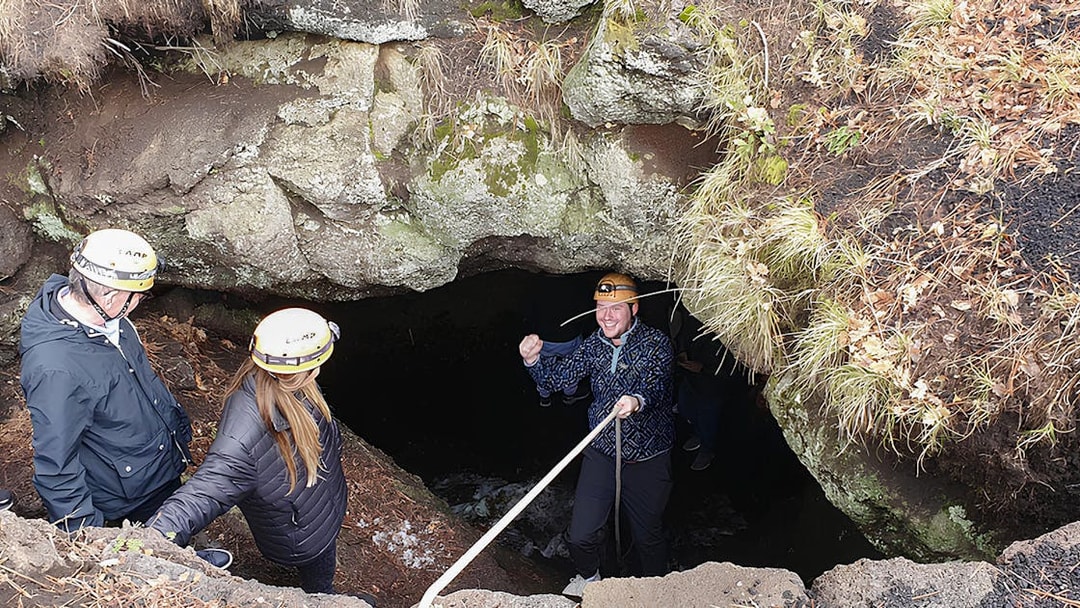 Tourists with helmets entering a lava cave guided by an instructor holding a rope.