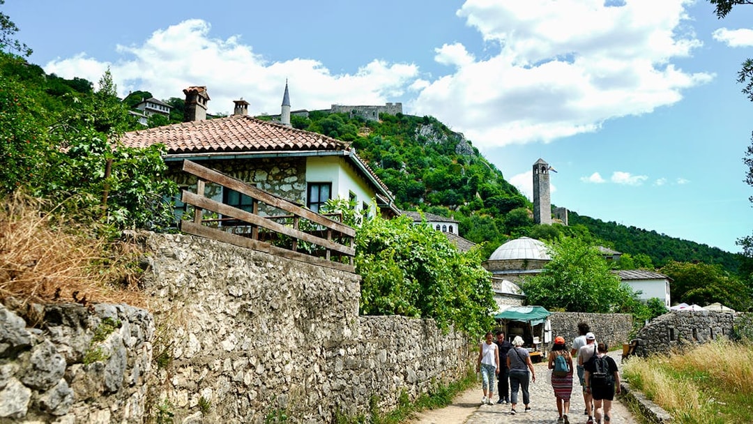 Tourists walking through historic Mostar with stone buildings and a hilltop fortress.