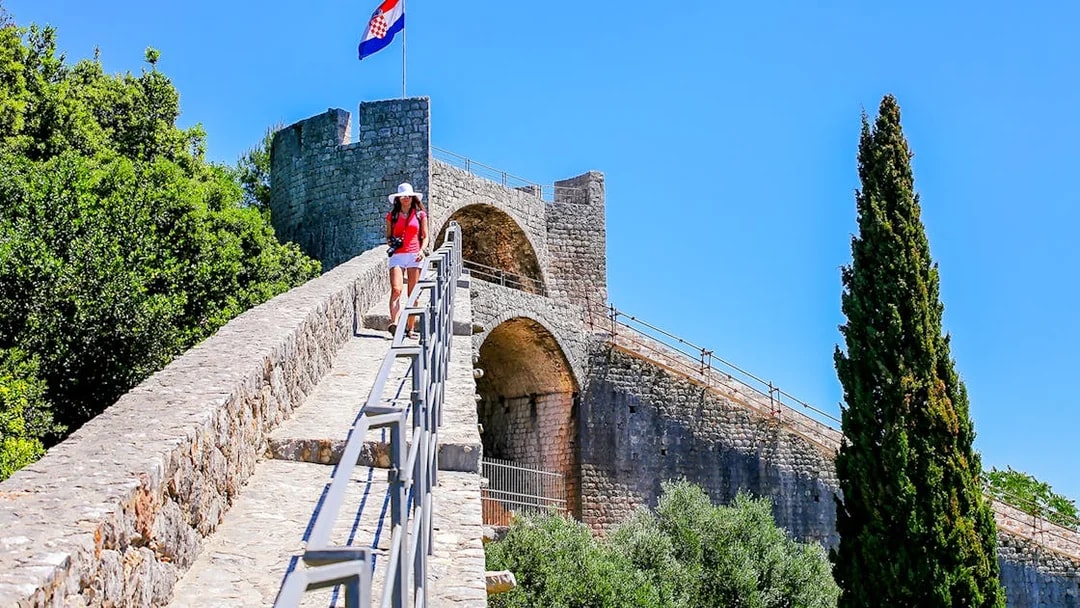 Young woman walking on the stone wall of Ston, Croatia, with a Croatian flag in the background.