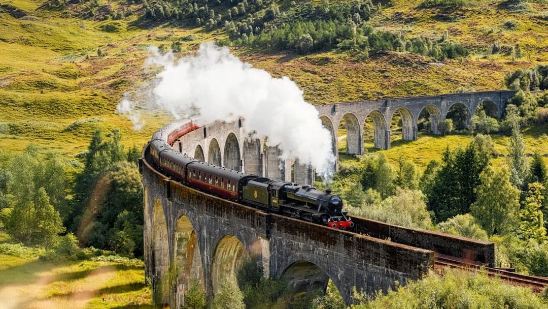 Steam train crossing Glenfinnan Viaduct in Scotland with lush green hills in the background.