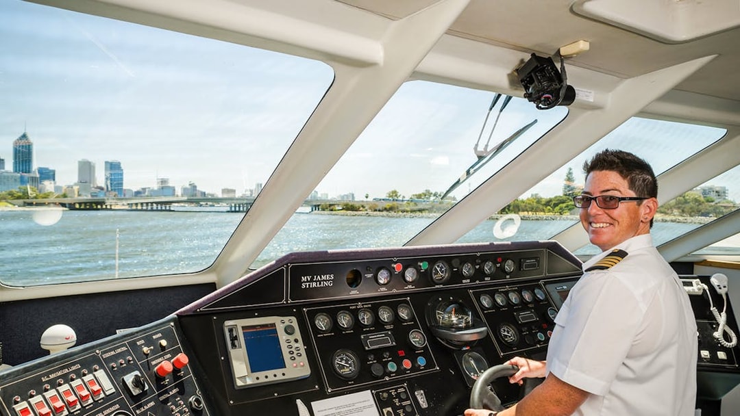 Pilot steering Fremantle Swan River Scenic Cruise with Perth skyline in view.