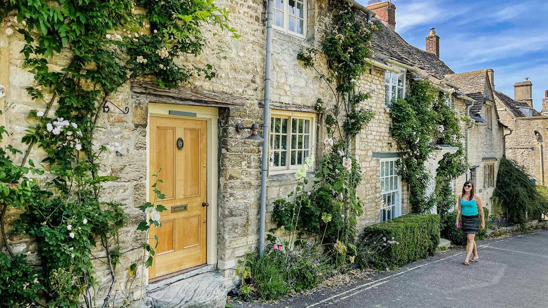 Cotswolds stone cottage with climbing plants and a person walking nearby.