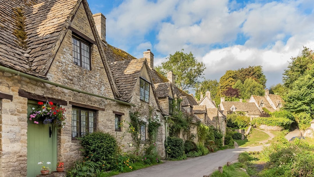 Cotswolds stone cottages along a village road, lush greenery, and blue sky.
