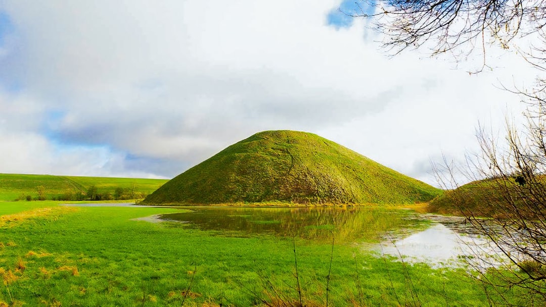 Silbury Hill in Avebury, England, surrounded by green fields and a reflective pond.