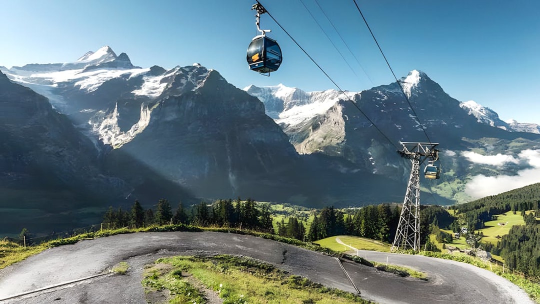 Cable cars over paths with snow-covered peaks and green valleys at Grindelwald First, Swiss Alps.