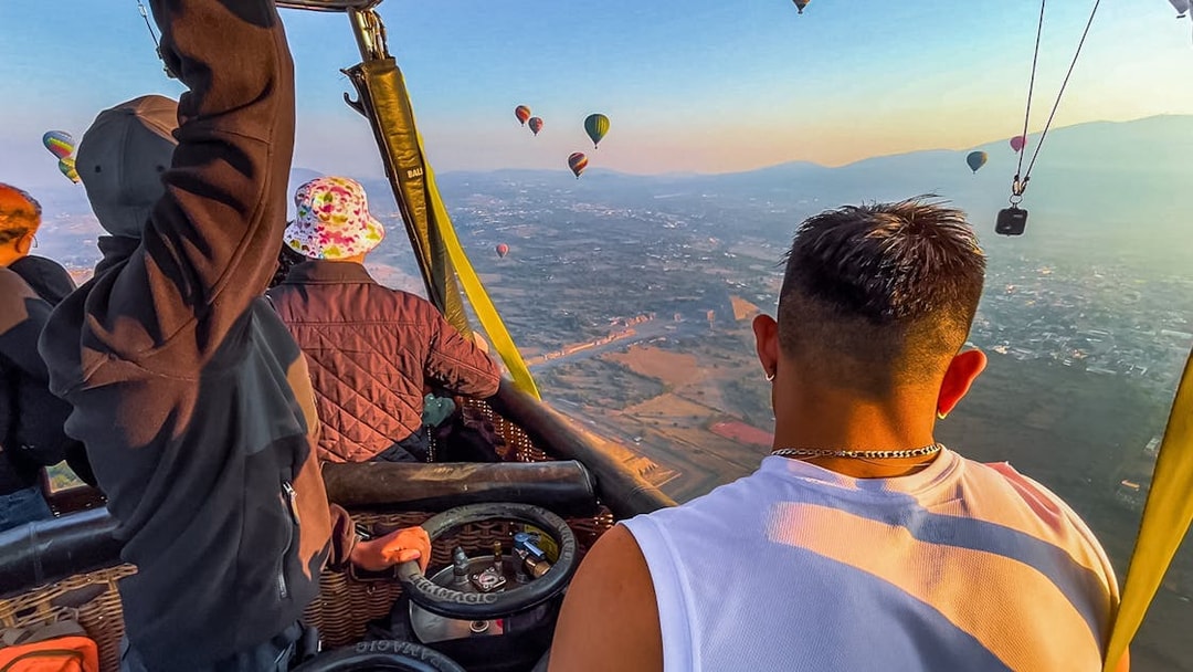 Passengers in a hot air balloon over Teotihuacan pyramids, Mexico, with other balloons in the sky.