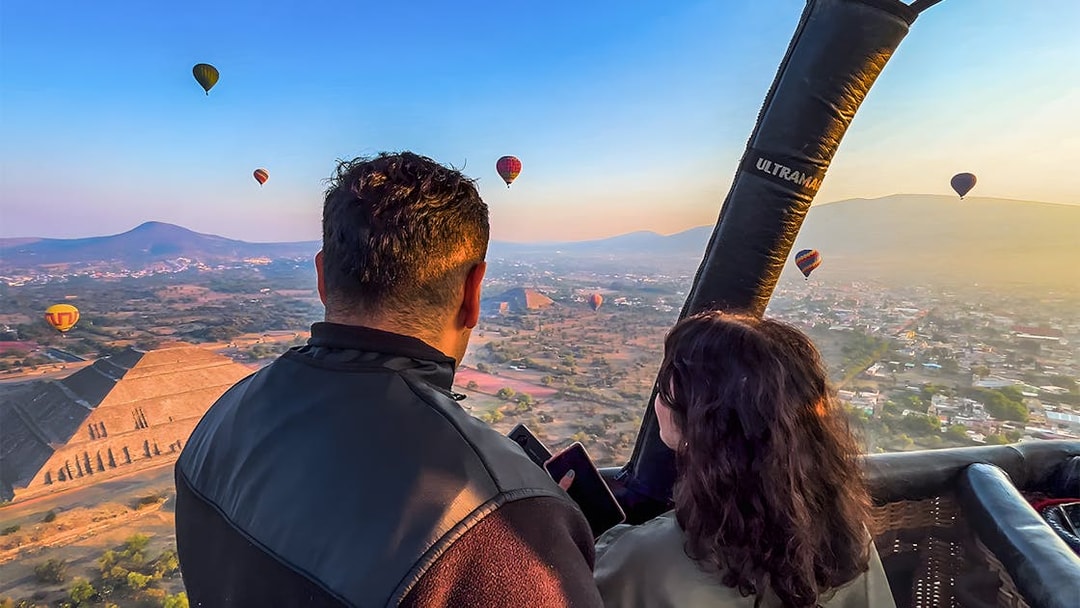 Passengers in hot air balloon over Teotihuacan pyramid, Mexico, with other balloons in the sky.