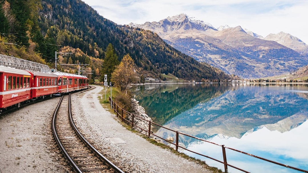 Red Bernina train traveling along a lake with mountain reflections near St. Moritz.