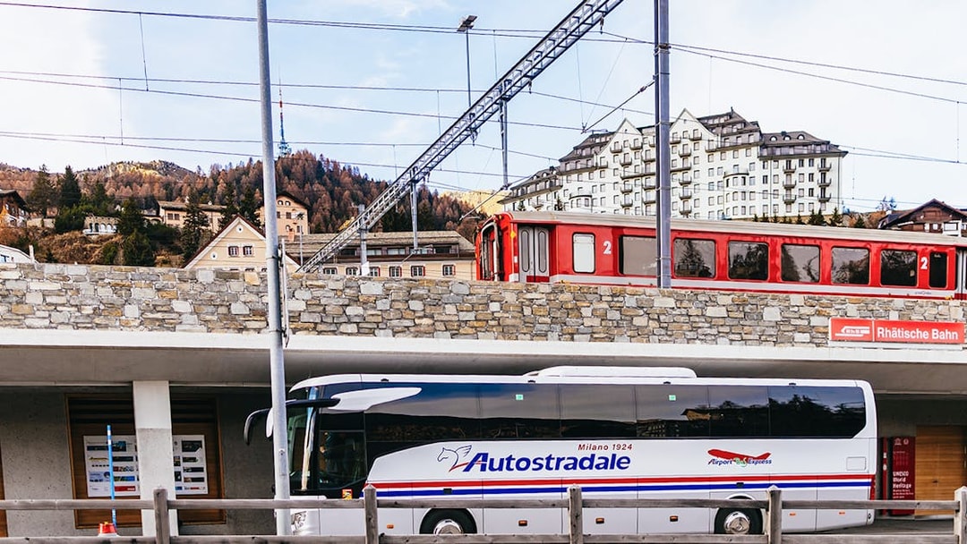 Bernina Red Train passing through St. Moritz station with a tour bus parked below.