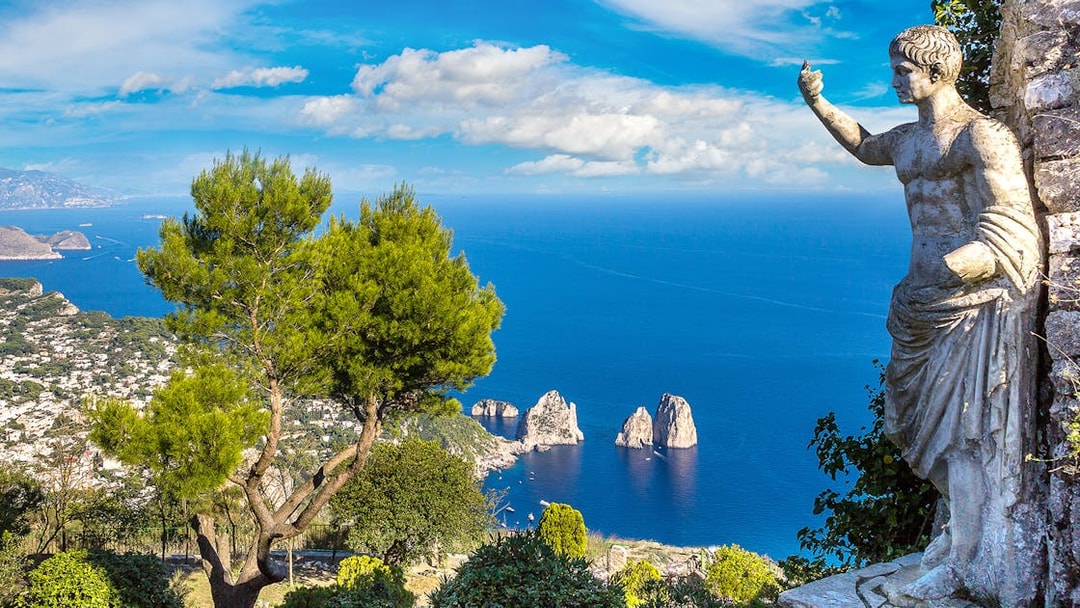 Capri coastline view with Faraglioni rocks and statue, seen from a hillside vantage point.