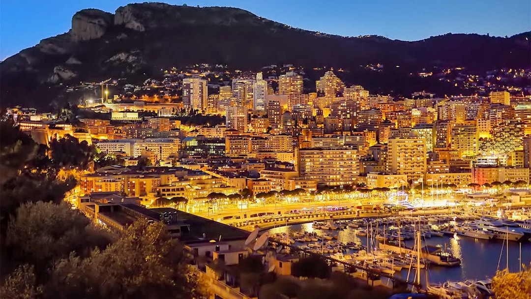 Monaco skyline at night with illuminated buildings and marina, viewed from Nice.