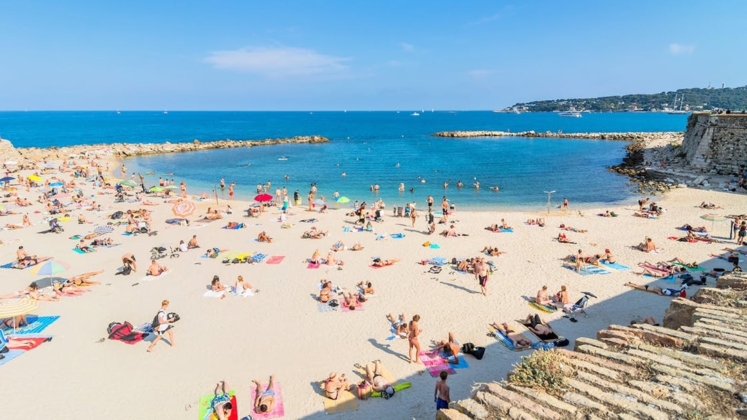 Beachgoers relaxing on a sandy beach in Antibes, France, with clear blue water and distant coastline.