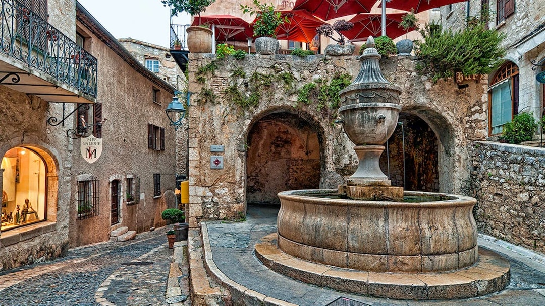 Stone fountain in a cobblestone street of Saint Paul de Vence, France, with rustic buildings and red umbrellas.