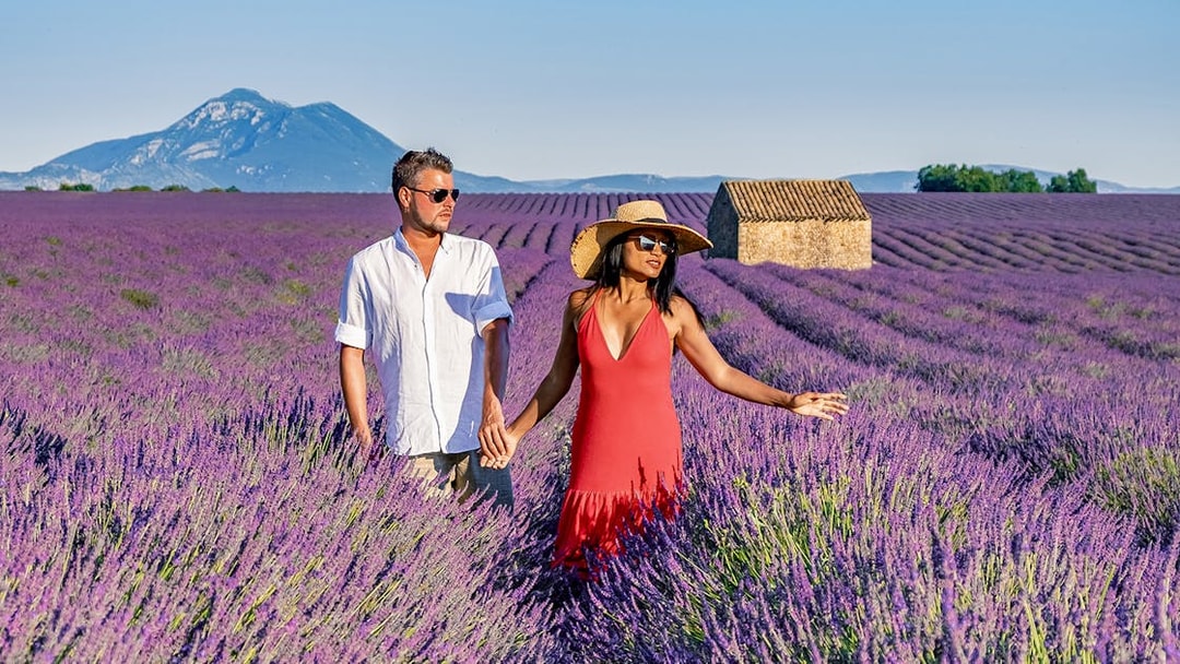 Couple walking through a lavender field in Provence with a mountain and stone house in the background.
