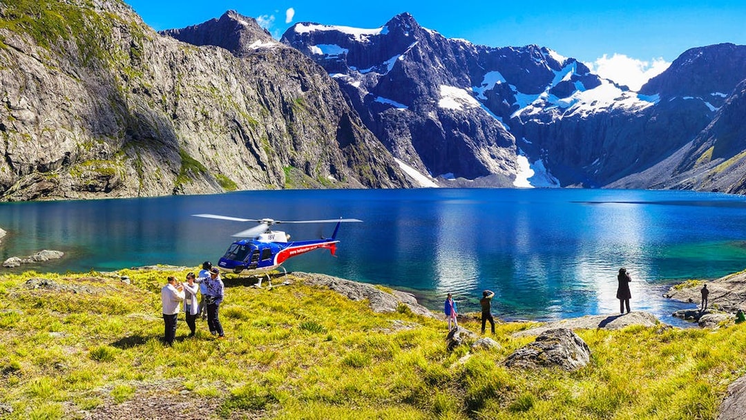 Helicopter landing by a lake in Milford Sound with people exploring the scenic mountain landscape.