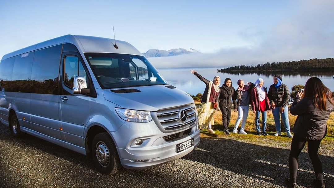 Group posing by tour van with scenic Milford Sound backdrop, Queenstown tour.