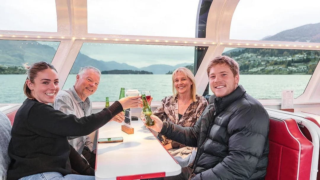 Passengers toast with drinks inside a cruise boat cabin, Queenstown scenery visible through the window.