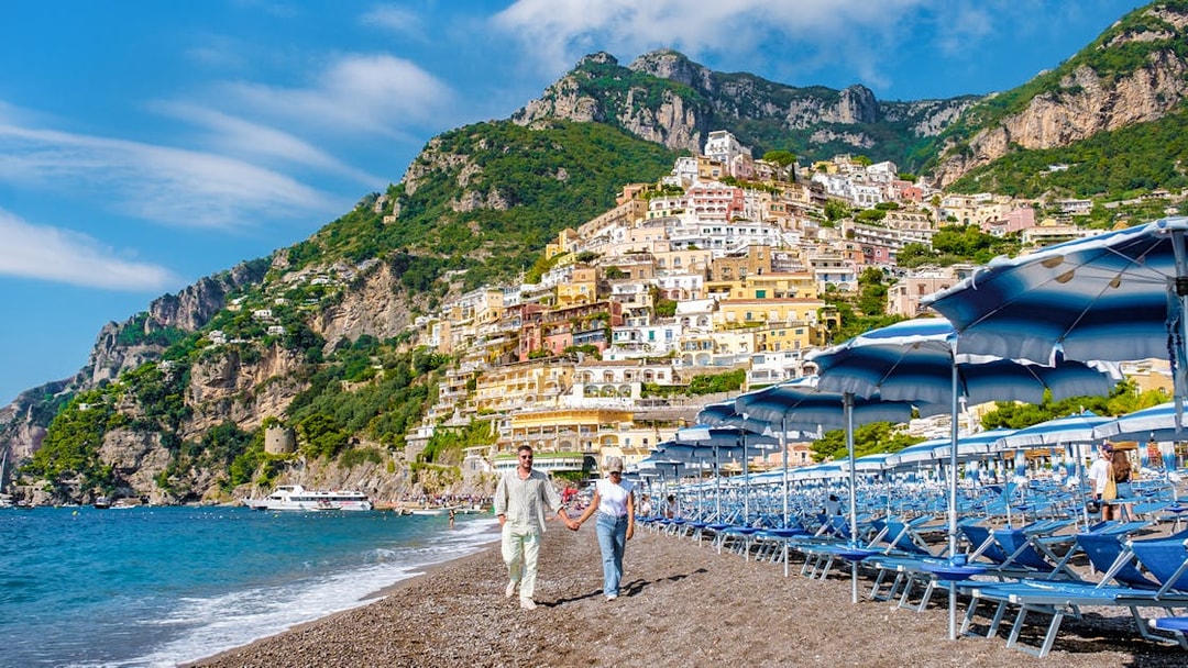 Couple walking on Positano beach with colorful hillside buildings, Amalfi Coast.