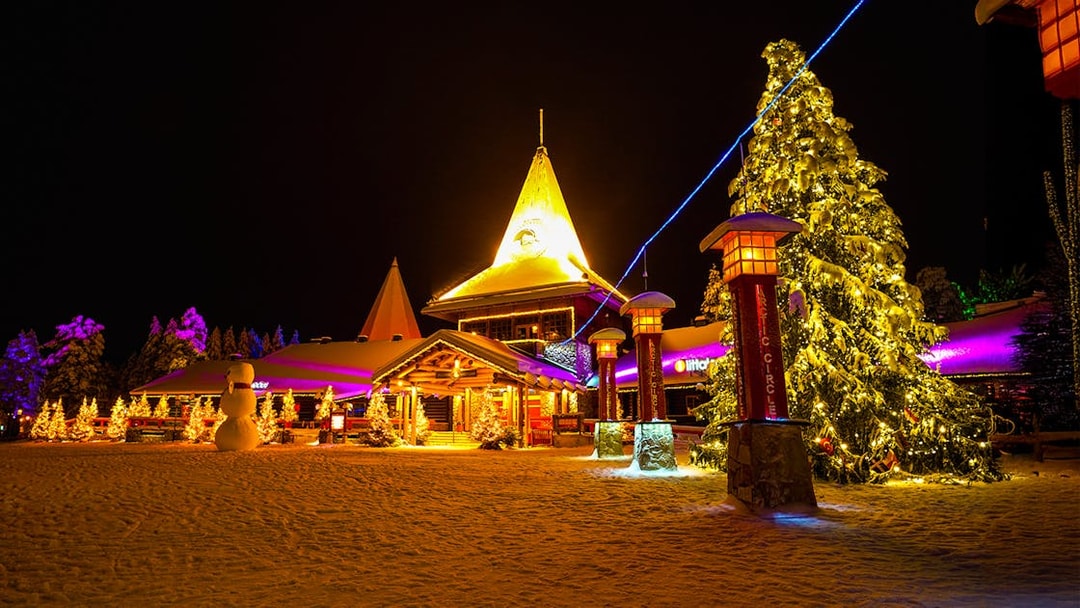 Santa Claus Village in Rovaniemi illuminated at night with snow-covered trees and festive lights.