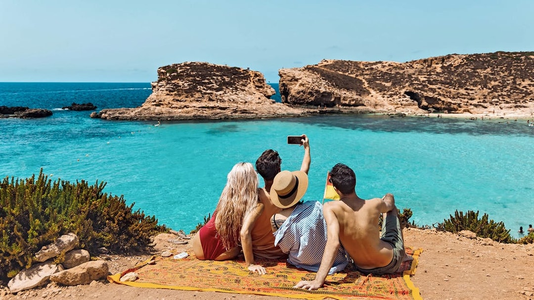 Friends taking a selfie at Blue Lagoon, Malta, with clear turquoise water in the background.