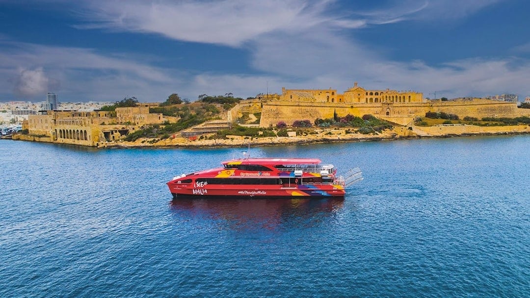 Red tour boat cruising near historic buildings in Valletta, Malta.