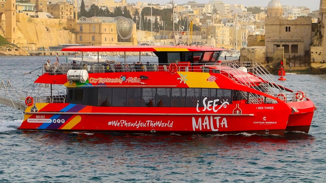 Red sightseeing boat cruising in Valletta harbor, Malta, with historic buildings in the background.