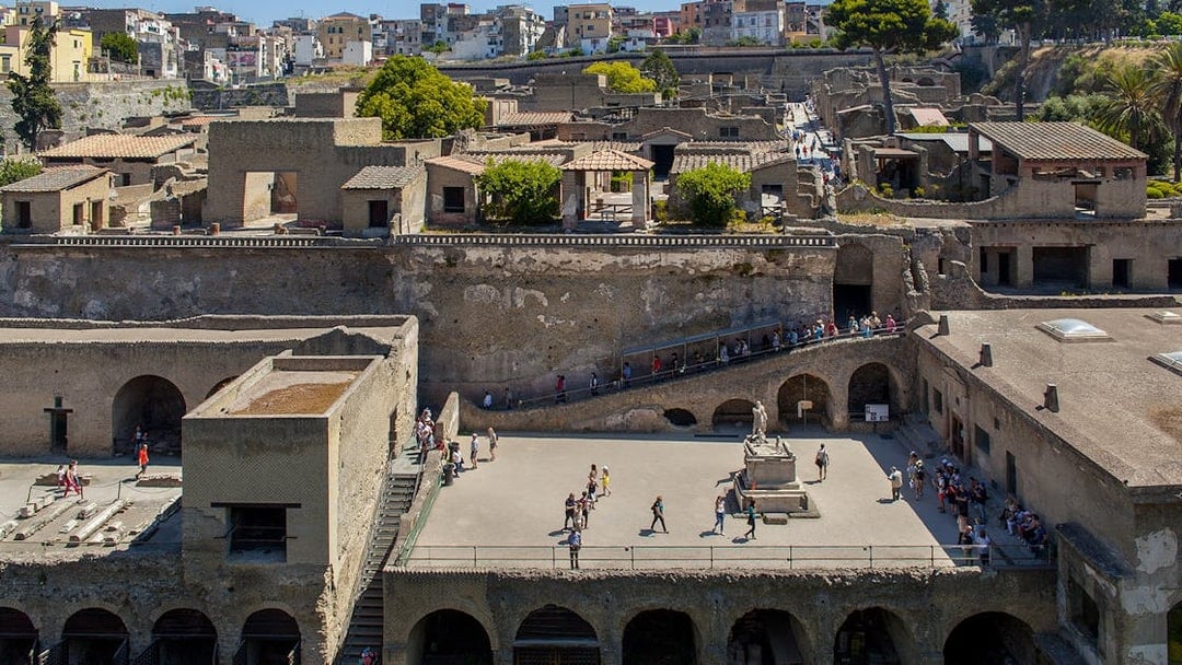 People walking through the ancient ruins of Herculaneum, Sorrento, with historic buildings in the background.