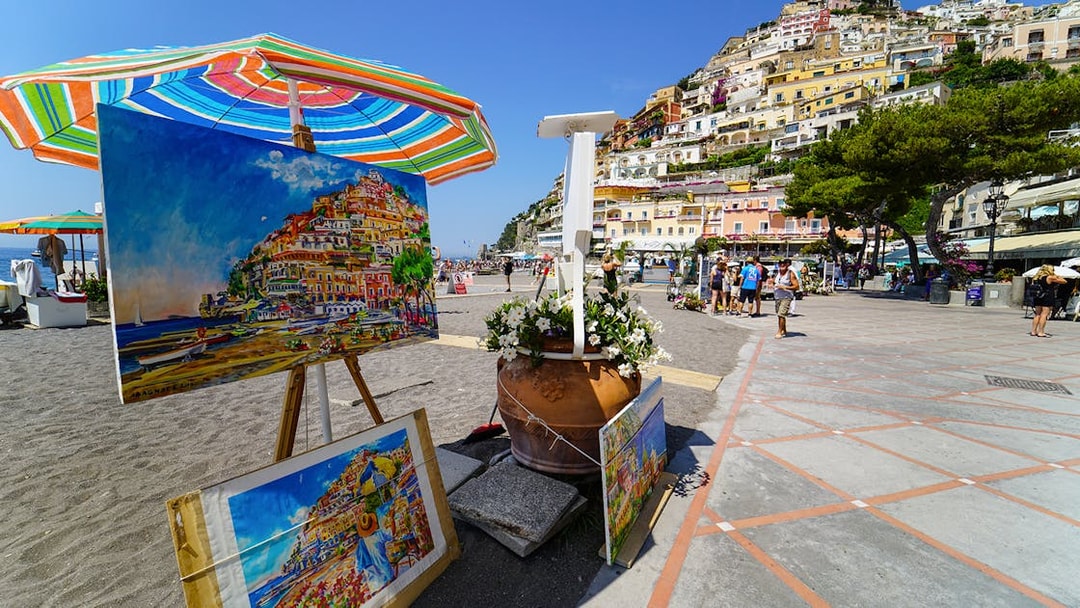 Colorful paintings of Positano displayed on a beach promenade, with hillside houses in the background.