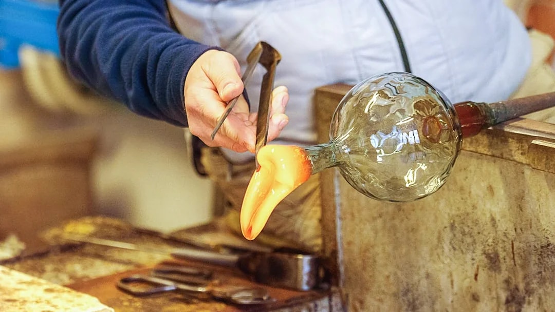 Glassblower shaping molten glass with tools in a workshop.