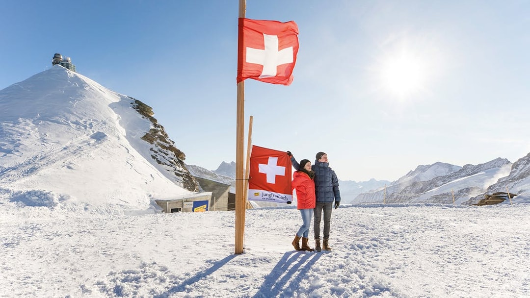 Couple standing by Swiss flags at snowy Jungfraujoch, with observatory and mountains in background.