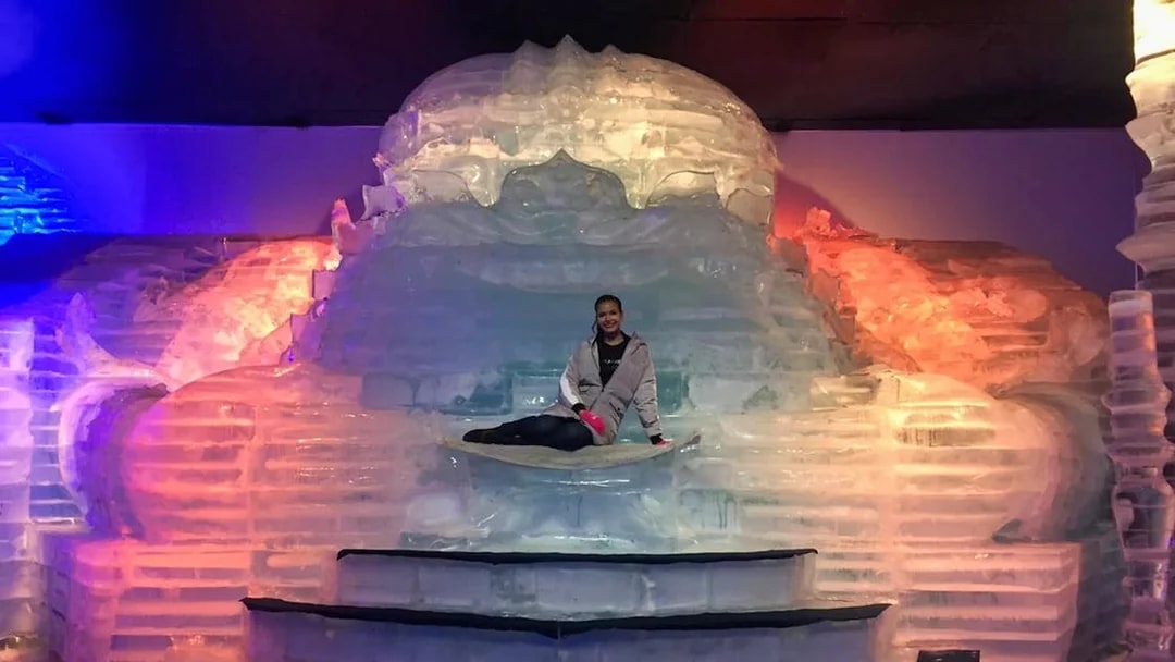 Person sitting on an ice sculpture at Frost Magical Ice of Siam, illuminated by colorful lights.