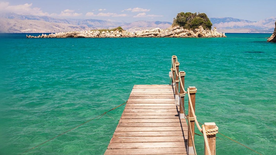 Wooden pier extending into turquoise sea with distant rocky islet, Corfu boat trip.