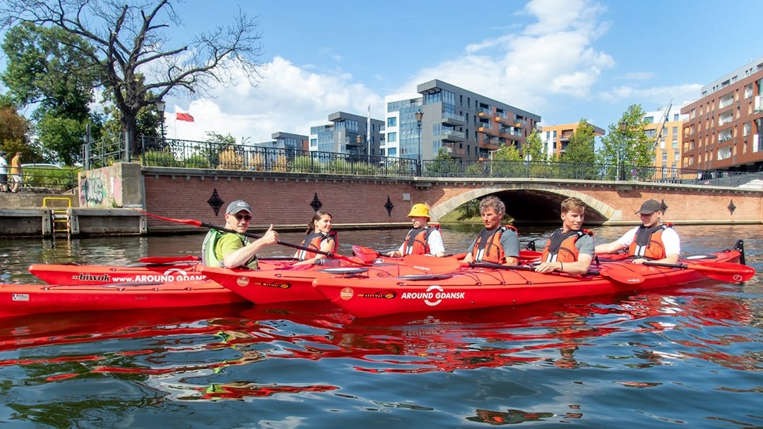 Kayakers paddling on a river in Gdansk with modern buildings and a bridge in the background.