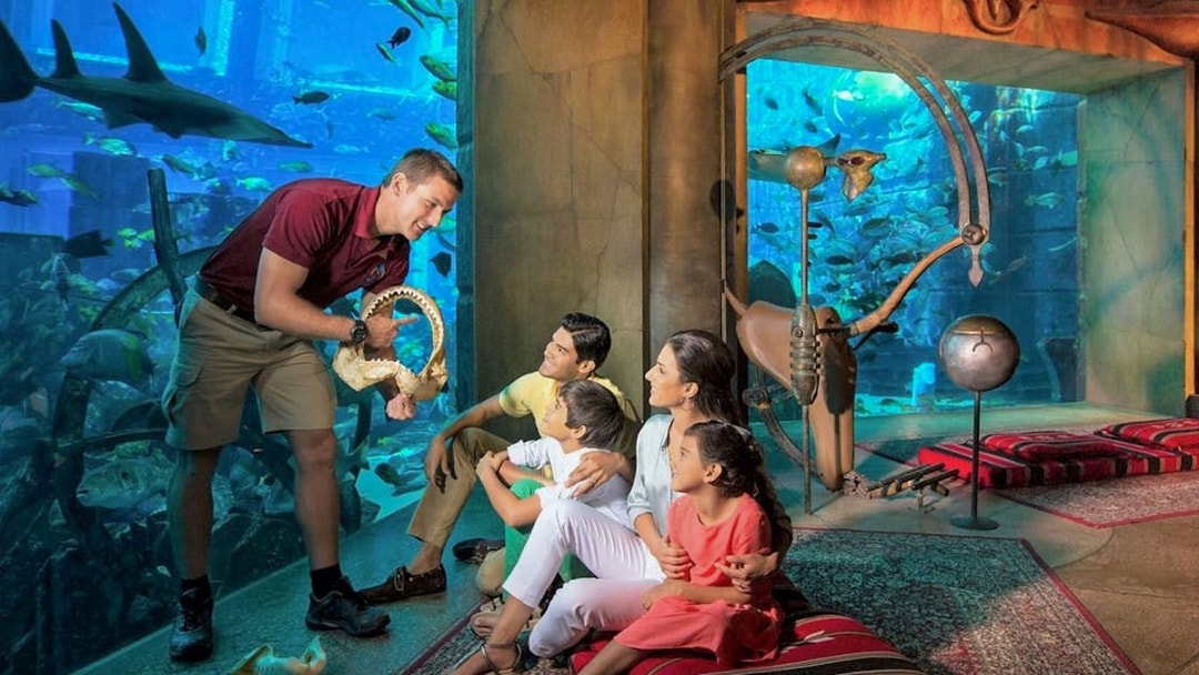 Family observing marine life at The Lost Chambers Aquarium, Dubai, with a guide showing a shark jaw.